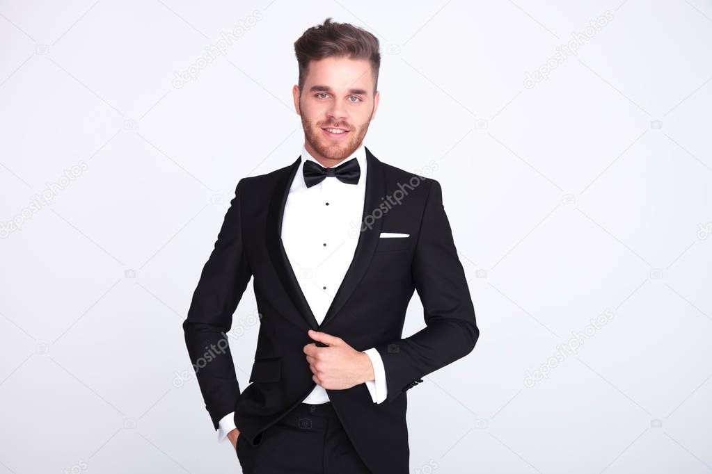 portrait of relaxed stylish man buttoning his black suit while standing on light grey background