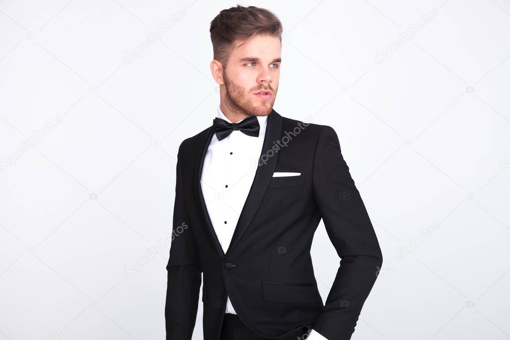 portrait of stylish curious man wearing black tuxeo and bowtie looking to side while standing on light grey background