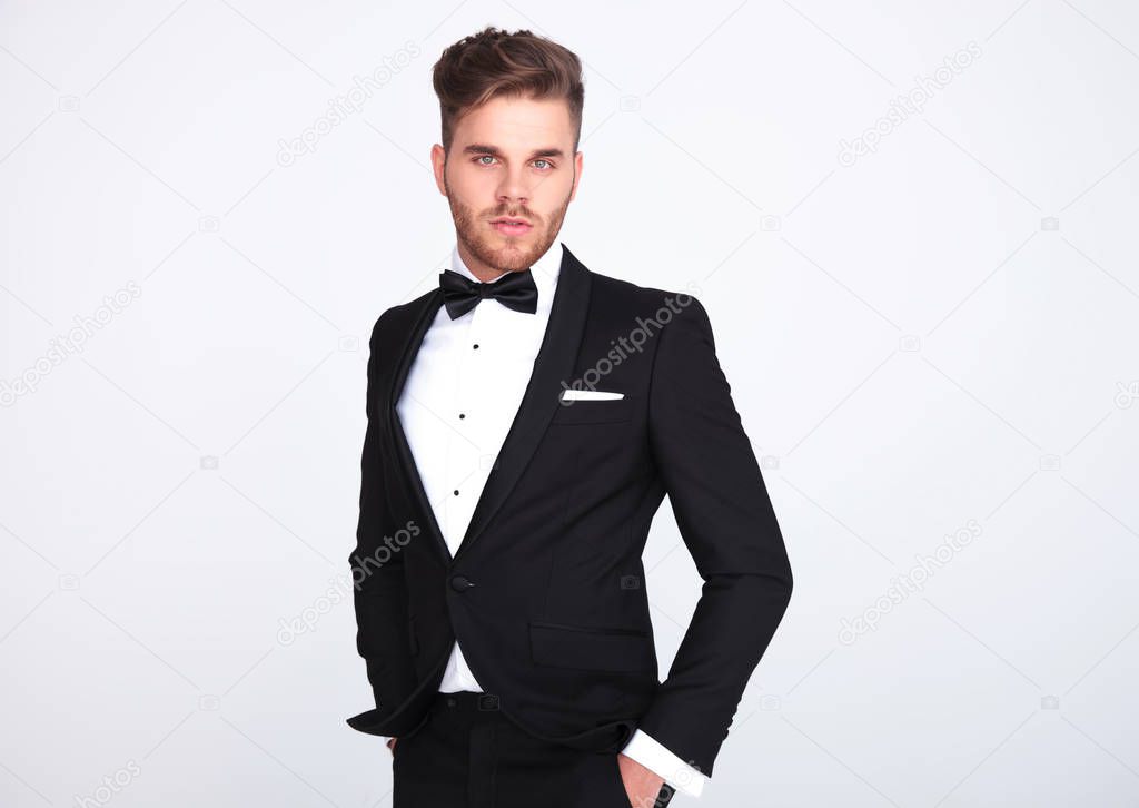 portrait of relaxed elegant man in black tuxedo standing on light grey background with hands in pockets