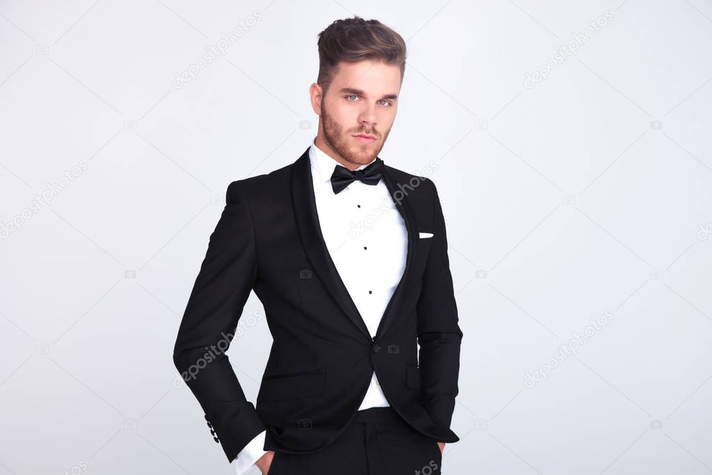 portrait of relaxed man in black tuxedo standing with hands in pockets on light grey background