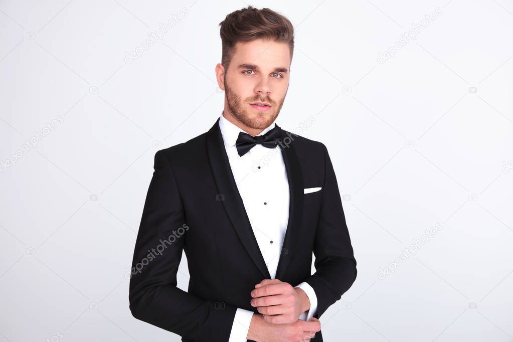 portrait of young elegant man in black tuxedo fixing his links while standing on light grey background