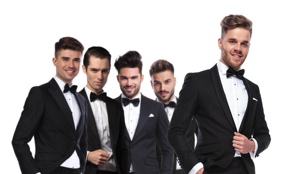 portrait of four men in tuxedoes in line with leader standing in front of them on white background