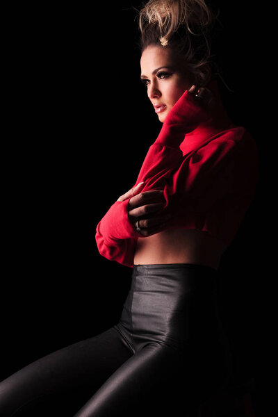 side view of sensual seated woman wearing red top and black leggings touching her neck on black background