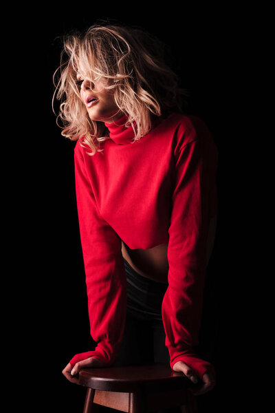 portrait of sexy woman in red top holding and leaning on chair and looking to side while standing on black background