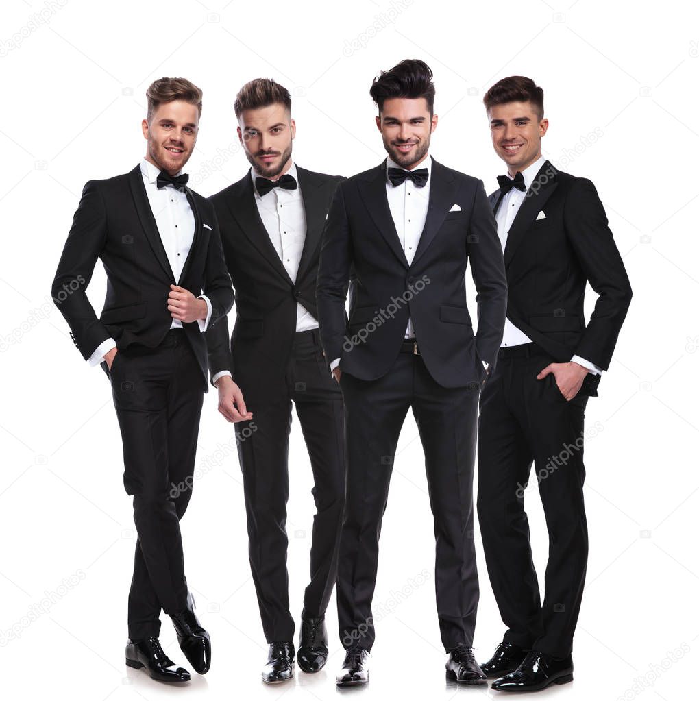 four elegant young men in tuxedos standing together on white background, looking relaxed. full body picture