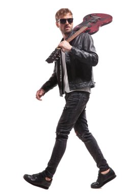 güneş gözlüğü ve omuz, tam vücut resim gitar ile beyaz arka plan üzerinde yürüme deri ceket giymiş yakışıklı rocker yan görünüm