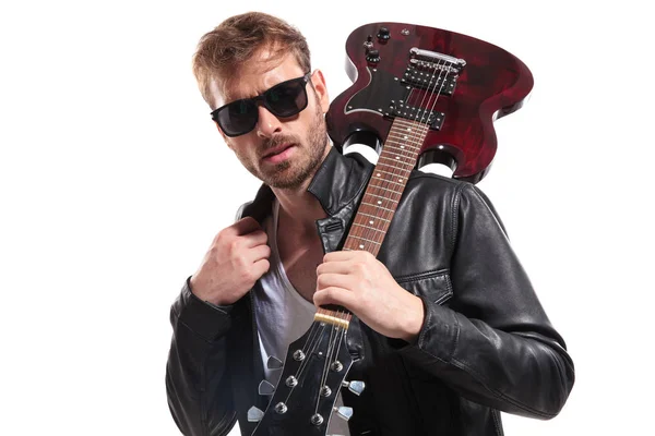 レザー ジャケット襟を押しながら彼のエレキギターの肩の上で白い背景の上に立ってサングラスを身に着けているセクシーなギタリストの肖像画 — ストック写真