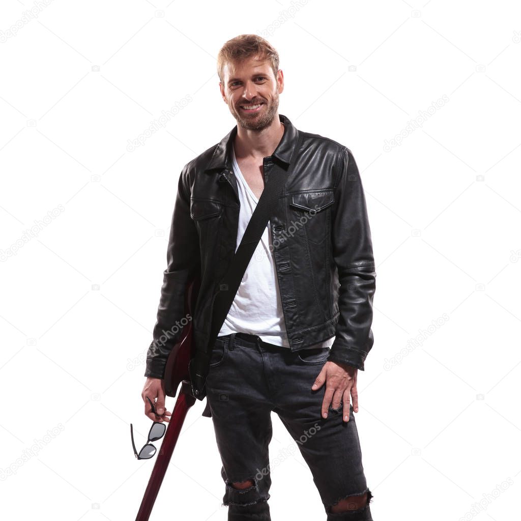 portrait of joyful young guitarist in leather jacket holding his sunglasses while standing on white background