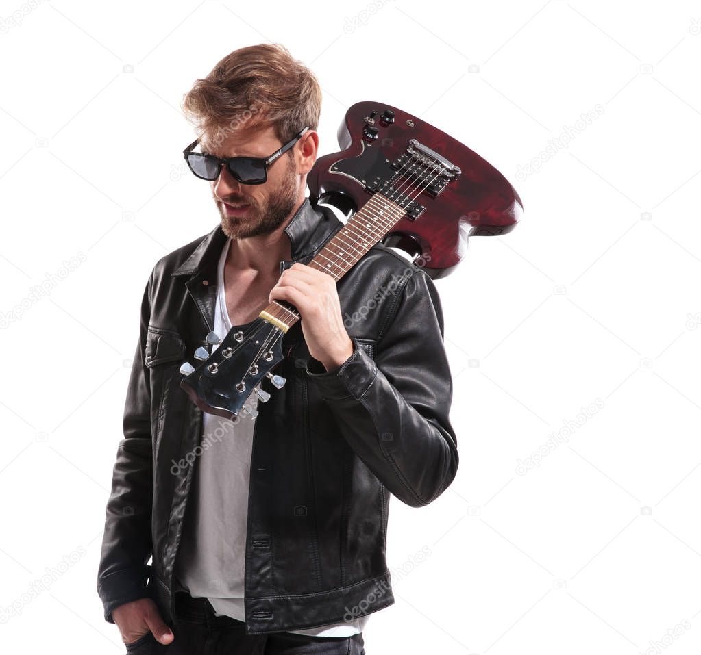 portrait of relaxed fashion man wearing sunglasses and leather jacket holding guitar on shoulder and looking down to side