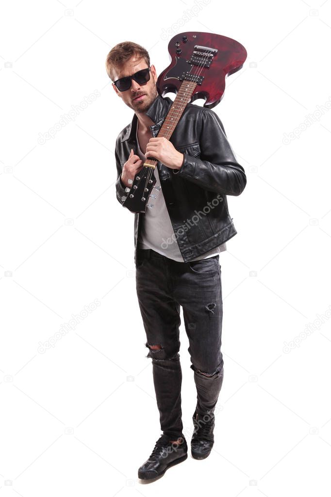 fashion man with guitar on shoulder walking on white background while holding leather jacket's collar, full body picture