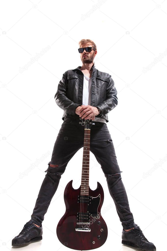 wide angle of dominant guitarist wearing black leather jacket and sunglasses holding his electric guitar while standing on white background
