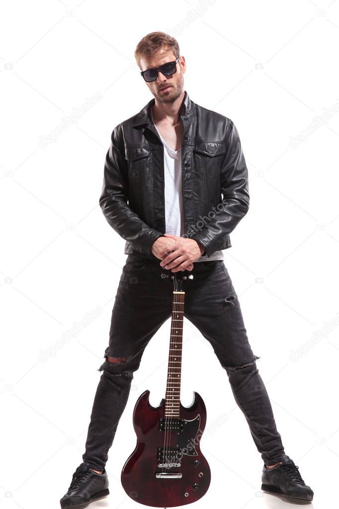 handsome rock star wearing sunglasses and leather jacket posing while holding his electric guitar and standing on white background