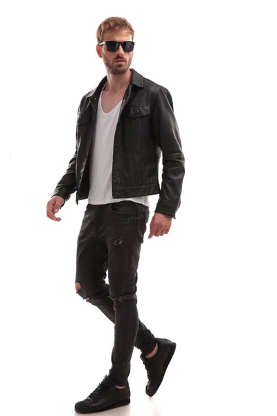 サングラスと黒い革のジャケットが白い背景のフルの長さの画像の上を歩くクールな若い男の側面図 — ストック写真
