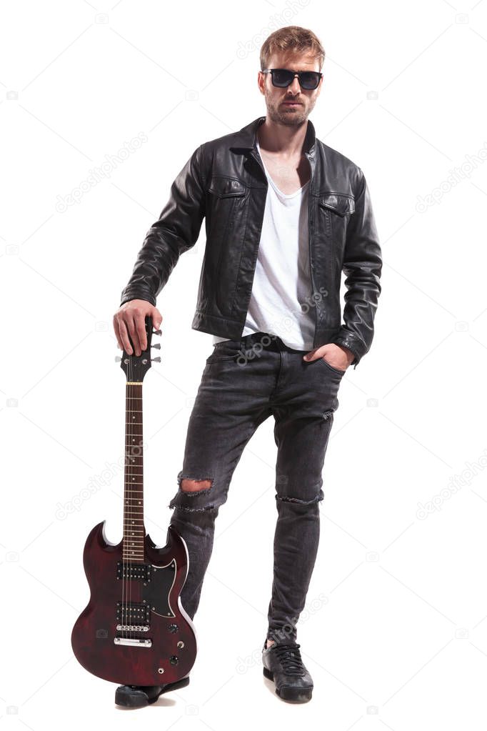 relaxed fashion man wearing black leather jacket and sunglasses posing with his electric guitar while standing on white background