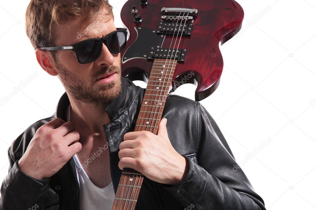 portrait of handsome man wearing sunglasses and leather jacket standing on white background with guitar on shoulder and looking to side