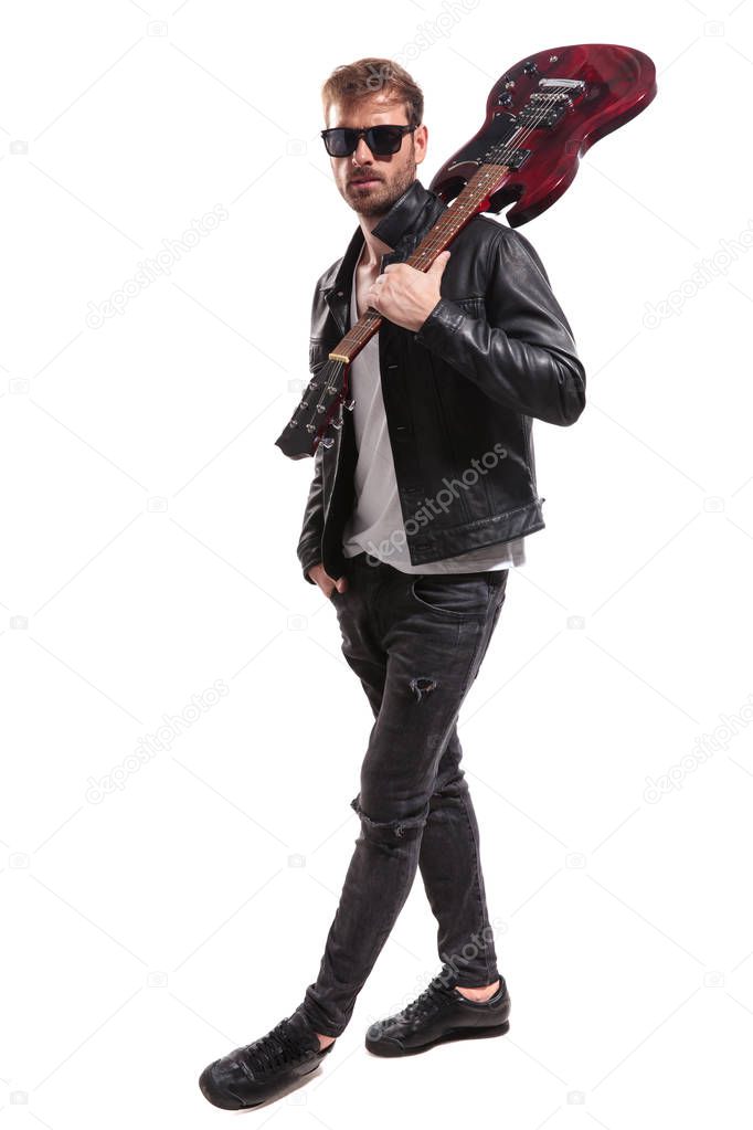 relaxed guitarist with sunglasses and leather jacket stepping to side while holding guitar on shoulder on white background