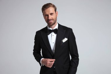 gülümseyen zarif adam onun smokin holding düğmesini ve kameraya gri arka plan üzerinde görünüyor