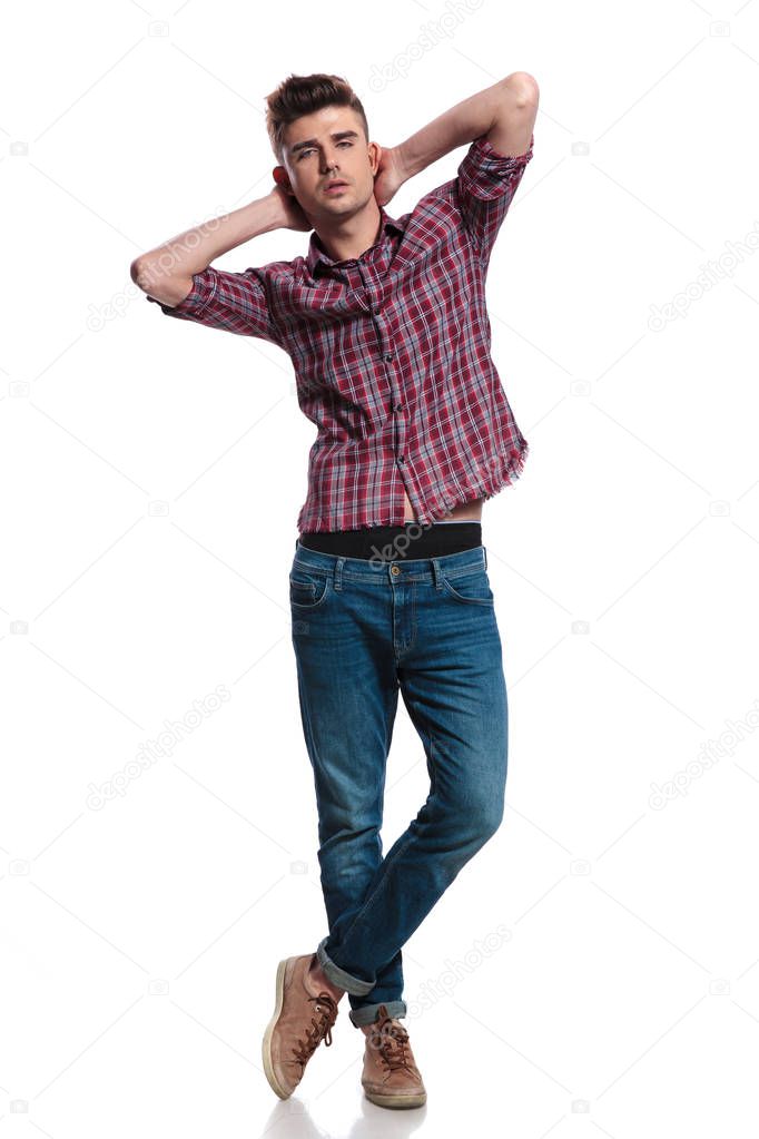 sensual man wearing shirt with plaid and jeans holds his neck while standing with legs crossed on white background, full length picture