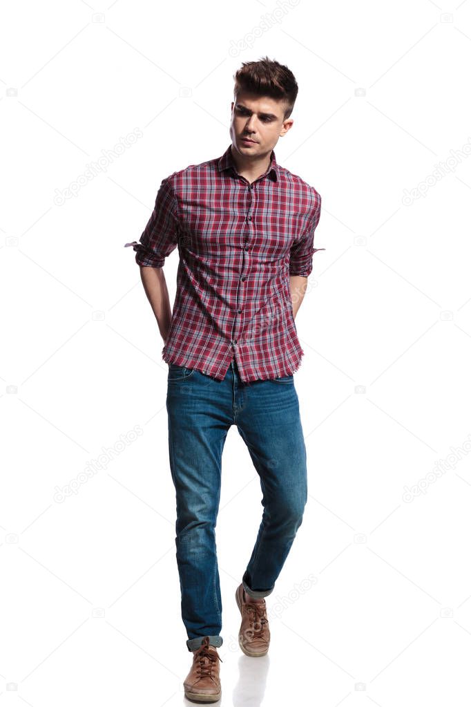 handsome casual man walking forward on white background looks down to side while holding hands in back pockets, full length picture