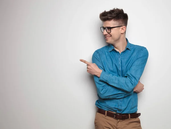 portrait of curious young man pointing and looking to side while standing on white background with arms folded