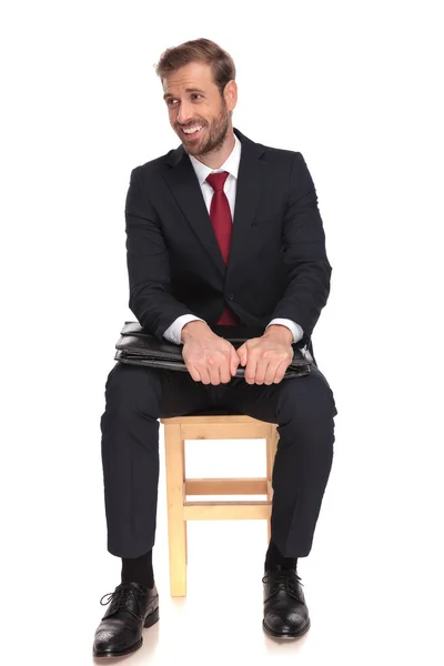 生意人坐在木椅子上 并在他的胳膊肘下举行公文包笑 看一边 在白色背景 — 图库照片