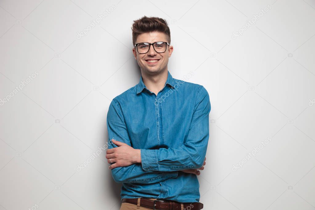 portrait of confident casual man wearing sunglasses smiling while standing on light grey background with hands folded