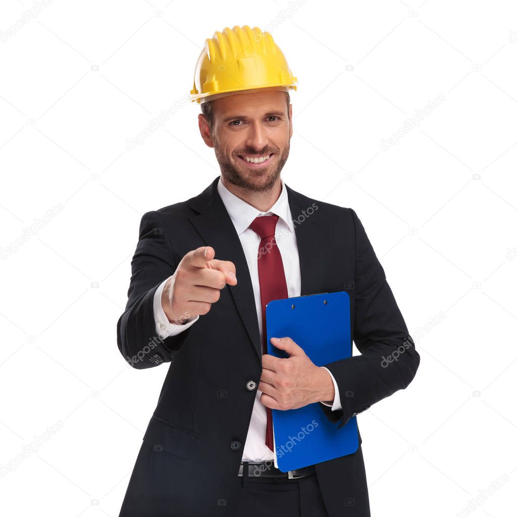 smiling businessman wearing protection helmet and holding clipboard points finger while standing on white background, portrait picture