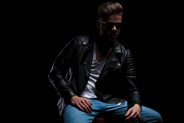 handsome young man wearing leather jacket and sunglasses sitting on wooden chair looks to side on black background