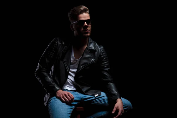 man wearing leather jacket and sunglasses sits on wooden chair and looks up to side on black background, portrait picture