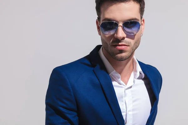 一个性感的年轻优雅的男人在蓝色西装和太阳镜看在灰色背景的相机特写镜头肖像 — 图库照片