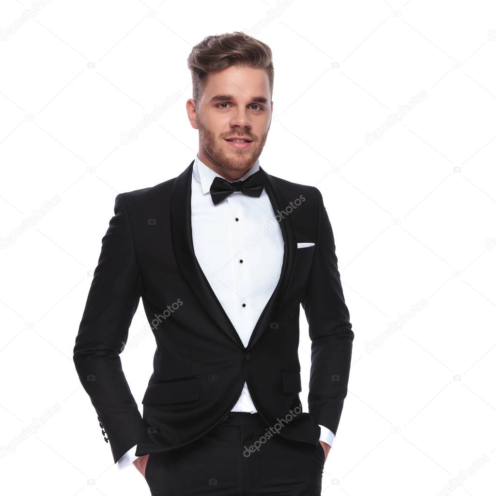 smiling elegant man in tuxedo standing with hands in pockets on white background