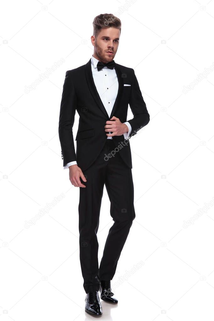 young groom opening his tuxedo while standing on white background and looks waya to side 