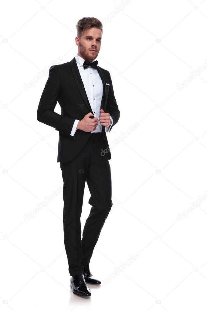 young elegant man puliing his tuxdo's collars while looking away from the camera on white background