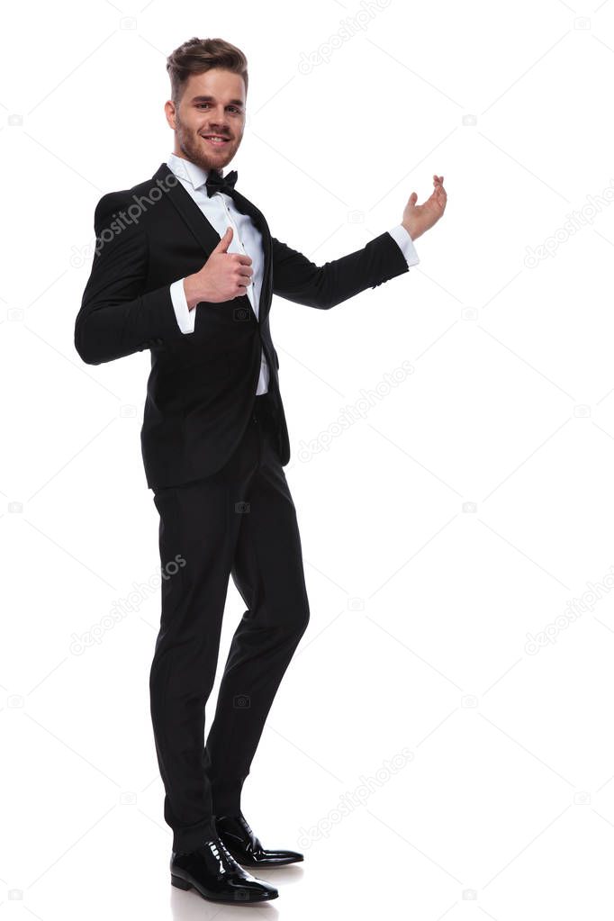happy elegant man in tuxedo presenting and making the ok thumbs up hand sign on white background