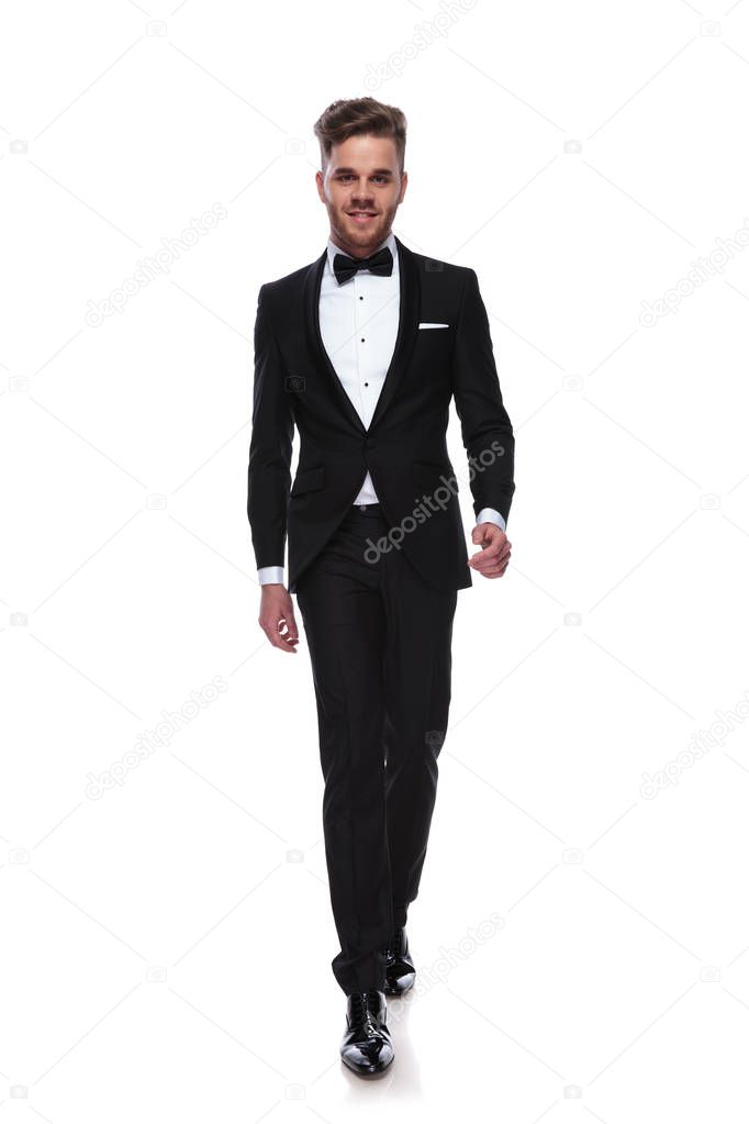 smiling young man in tuxedo and bow tie is walking forward on white background