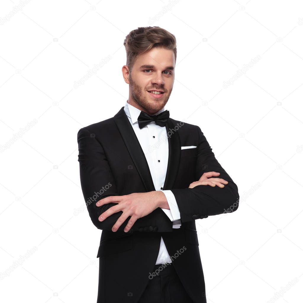 smiling elegant man in tuxedo standing with hands crossed on white background