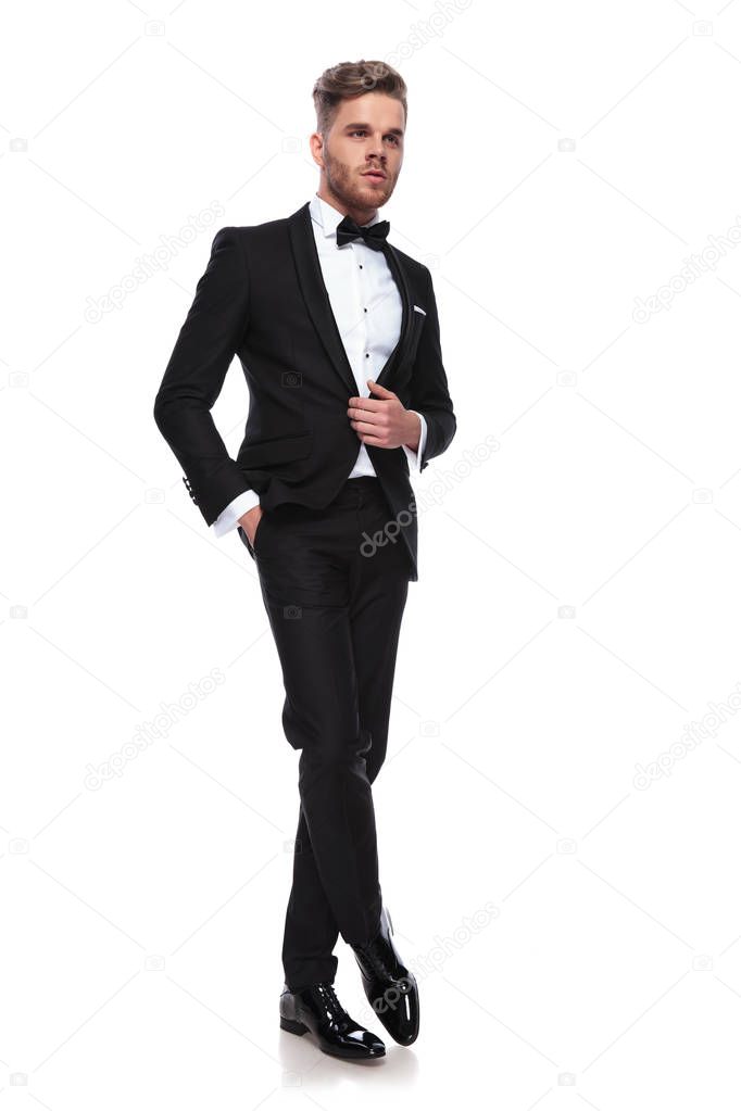 elegant man in tuxedo standing with legs crossed and holding his coat's button on white background
