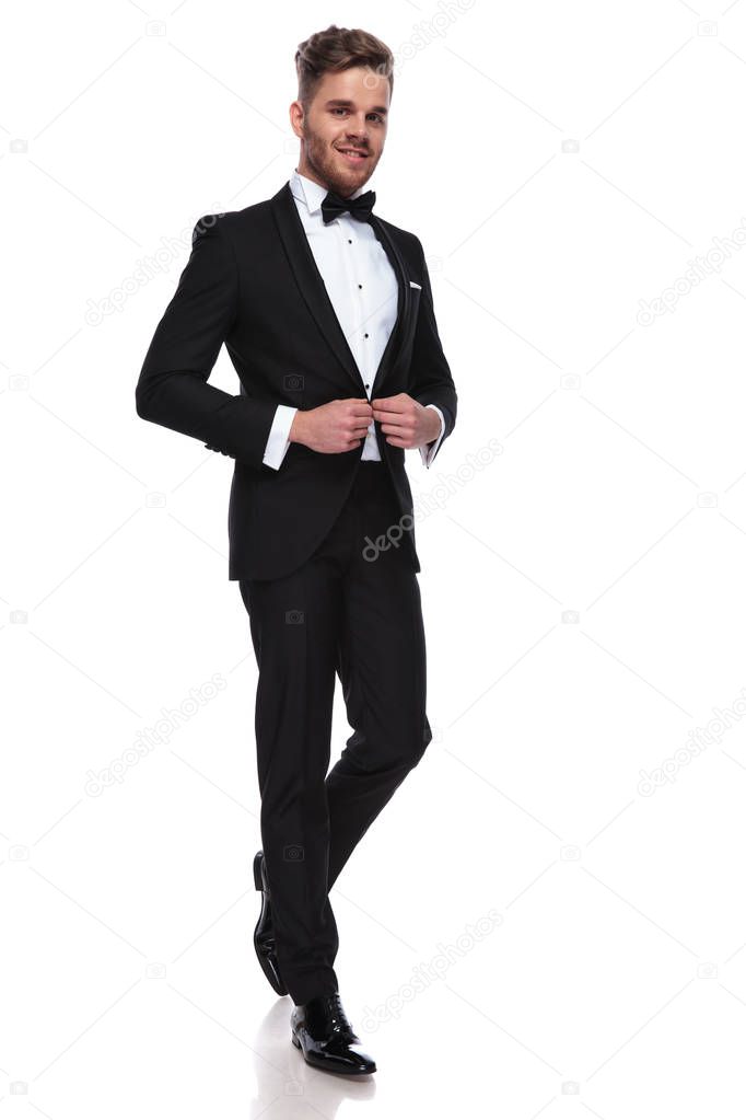 smiling young groom buttoning his tuxedo on white background