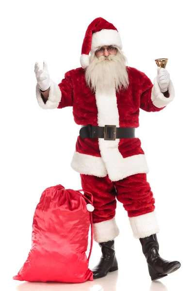 サンタ クロース彼の足に近い袋と白い背景の上に立って彼のベルが鳴ると歓迎のジェスチャー 全身画像 — ストック写真