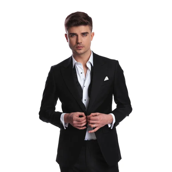 黒のタキシードを着て魅力的な若い男が白い背景の上に立っている間彼のスーツをボタンします — ストック写真