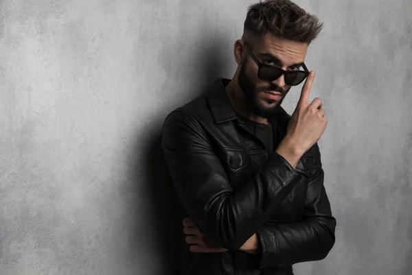 灰色の壁にもたれながらサングラスに触れる革ジャケットでカジュアルな男性の肖像画 — ストック写真