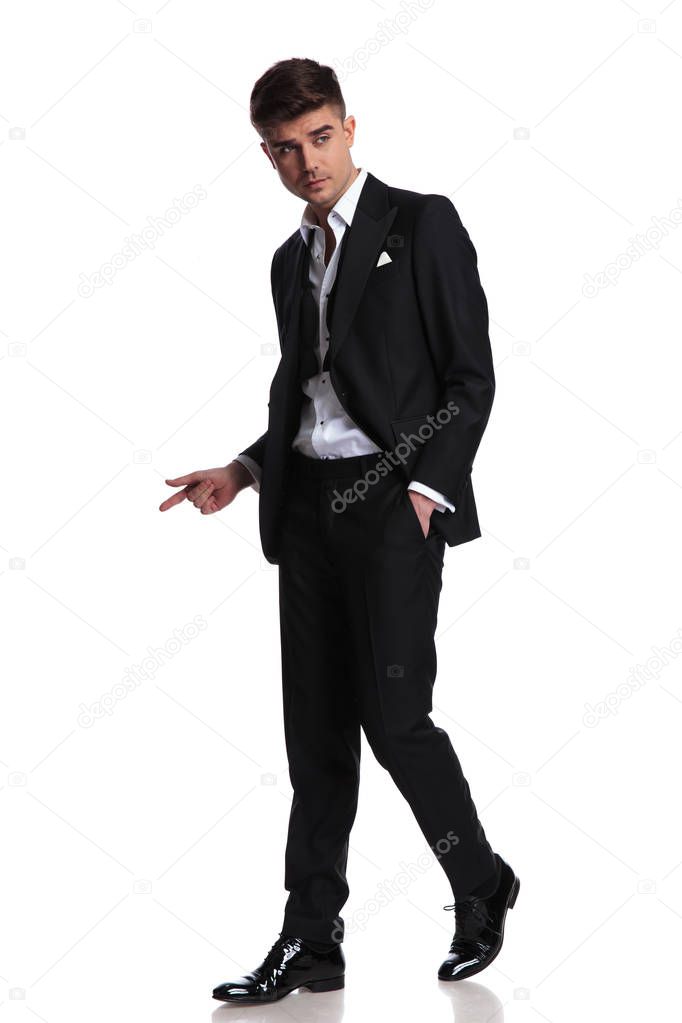 relaxed man in black tuxedo walks on white background to side and points to side