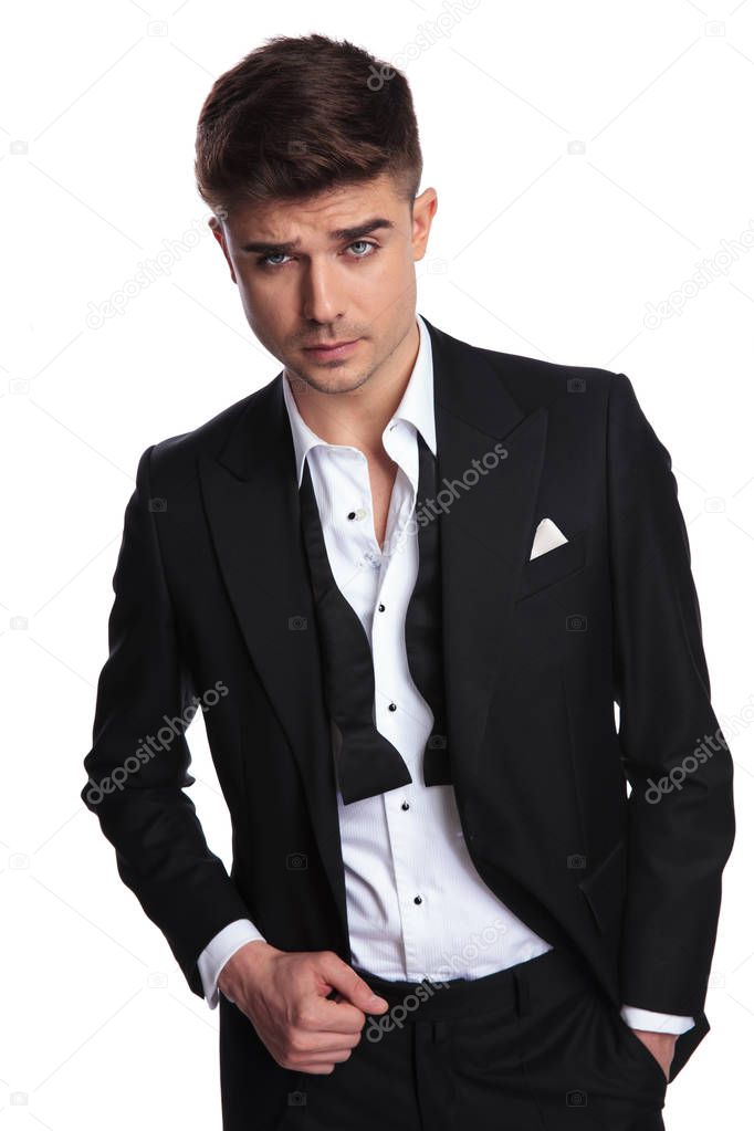 portrait of relaxed man in tuxedo looking up to side while standing on white background and holding tuxedo collar