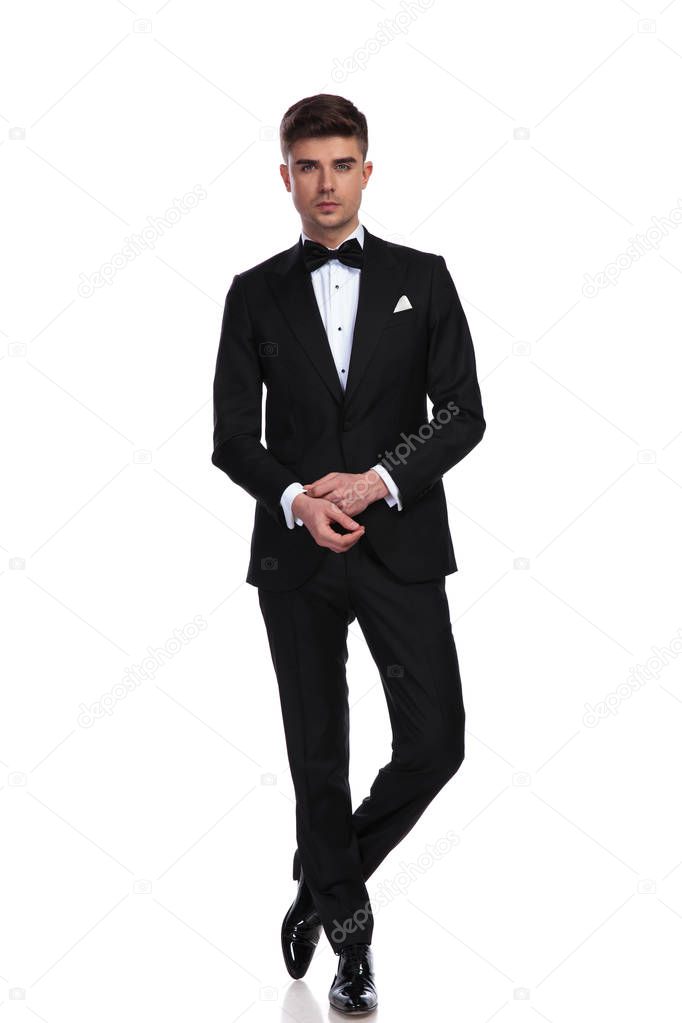 attractive groom fixing cuffs while standing on white background with legs crossed, full length picture