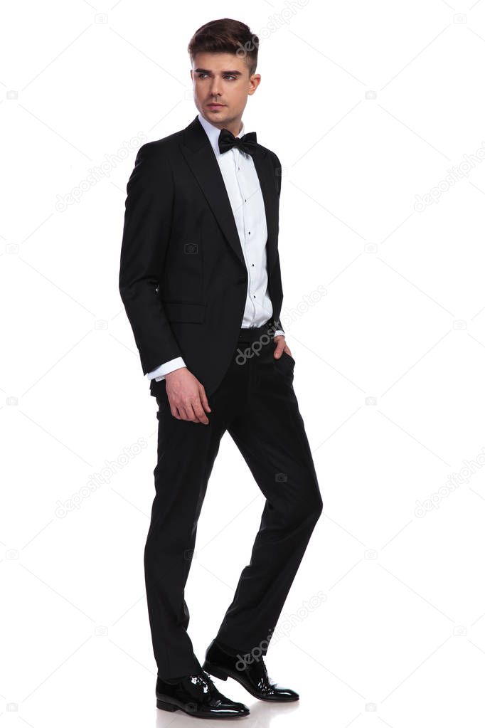 side view of relaxed businessman in tuxedo looking behind while standing on white background with a hand in pocket, full length picture