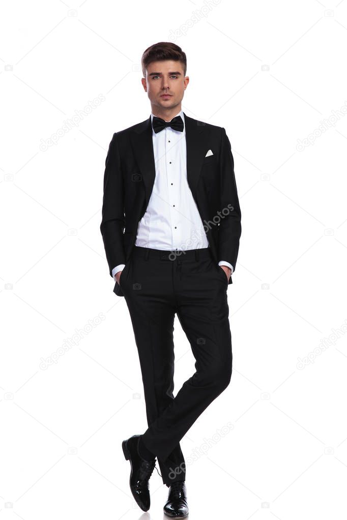 young groom in black tuxedo standing with hands in pockets on white background with legs crossed