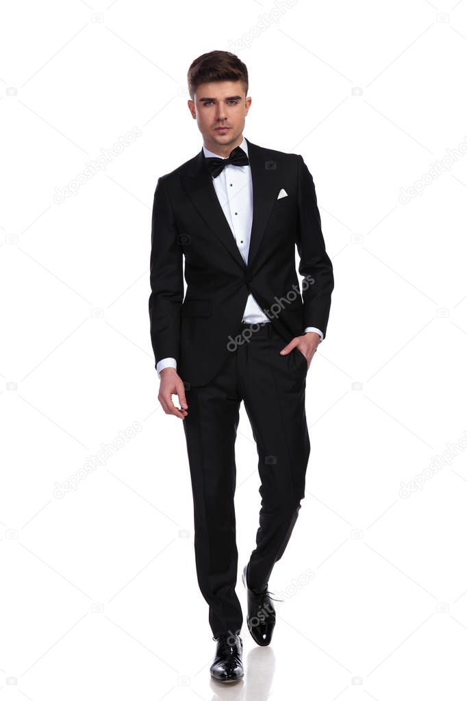relaxed groom in black tuxedo walks forward with hand in pocket on white background