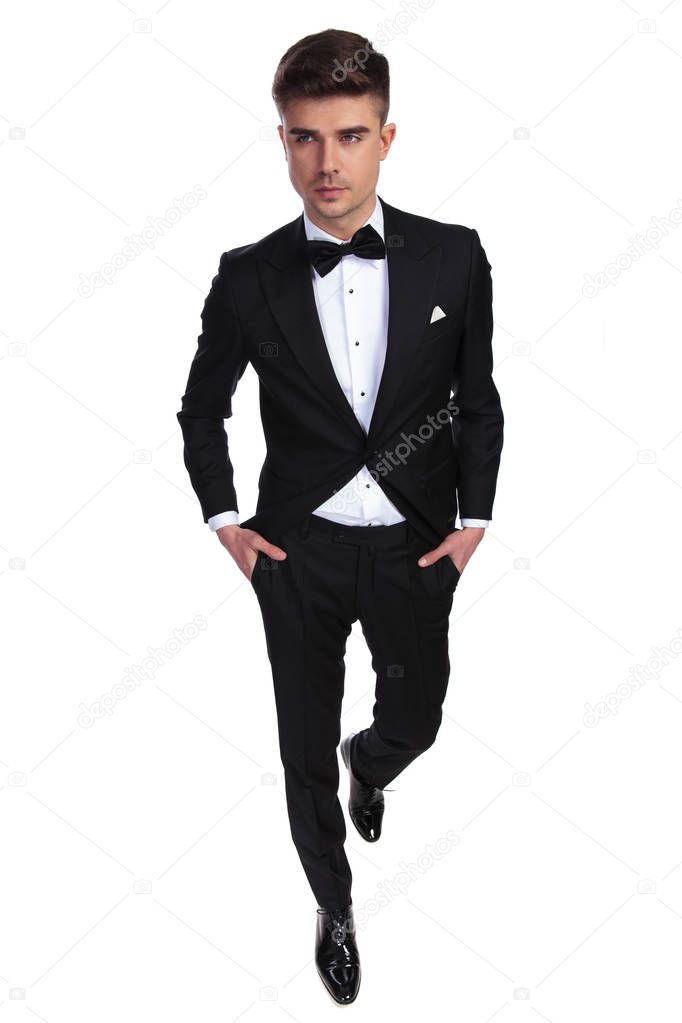 relaxed businessman in black tuxedo moves forward on white background and looks to side