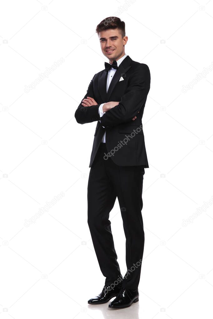 smiling groom in black tuxedo standing on white background with arms folded, full body picture