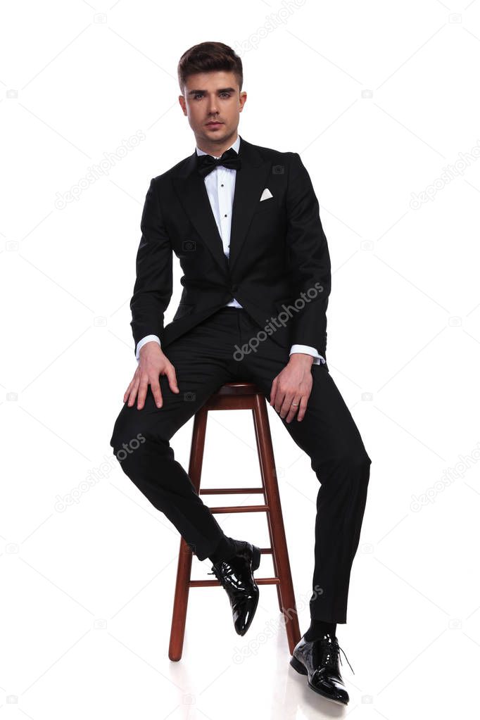 attractive groom in black tuxedo sitting on a wooden chair on white background